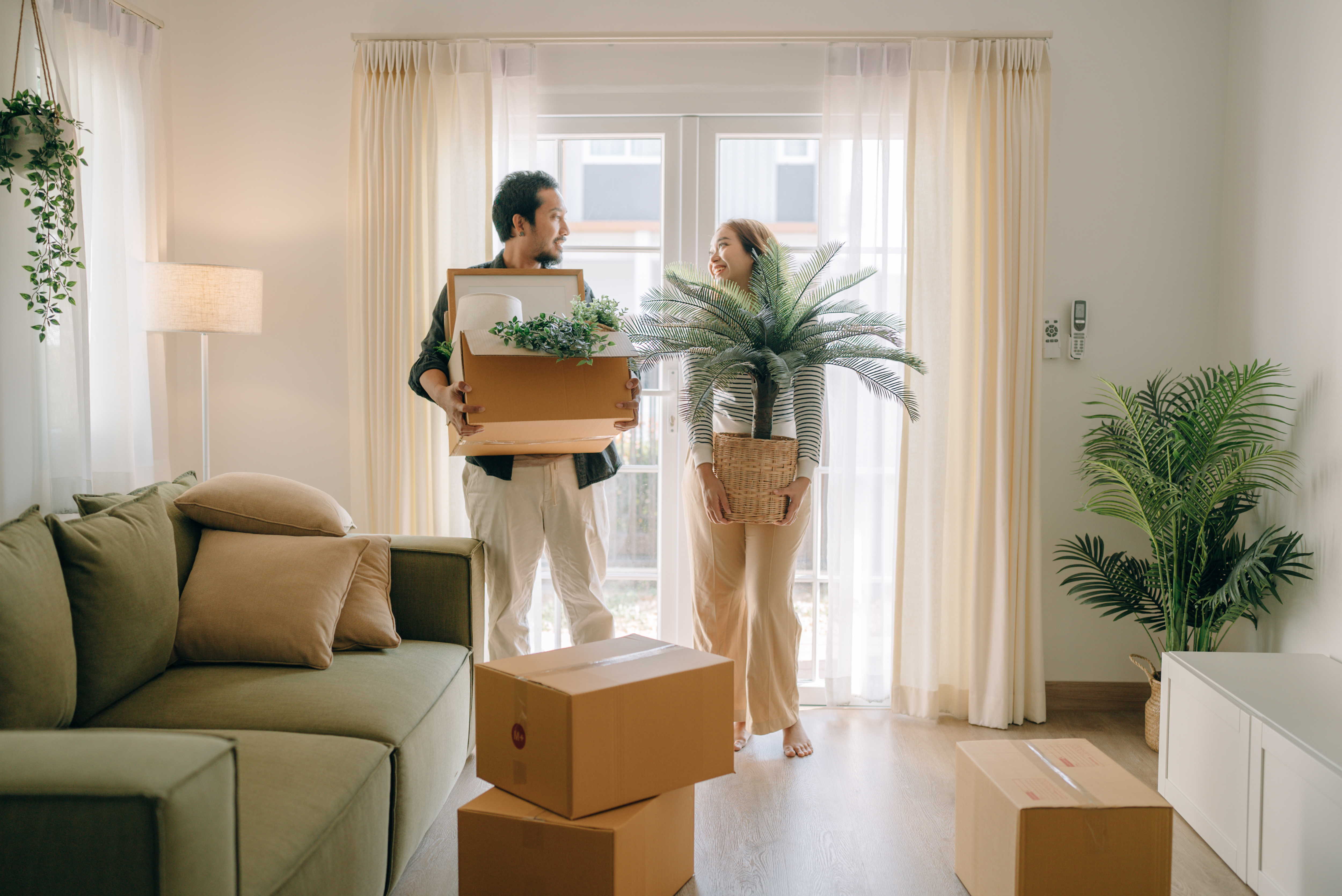 Two people stand in an apartment holding houseplants